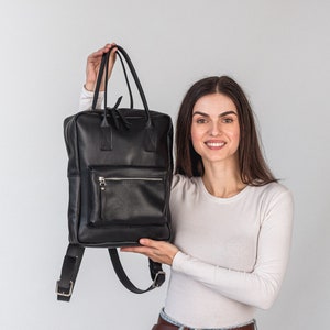 以下が含まれることがあります： 女性が、ジッパー付きポケットとシルバーの金具が付いた黒い革のバックパックを持っています。彼女は白い長袖シャツと、茶色のベルトが付いた青いジーンズを着用しています。