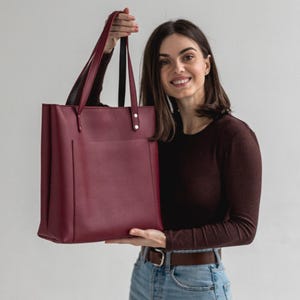 Peut inclure: Un sac cabas bordeaux profond de forme rectangulaire structurée, doté de deux longues anses fixées par des rivets métalliques argentés et d'une grande poche extérieure plaquée sur le panneau avant, avec une finition lisse et mate.