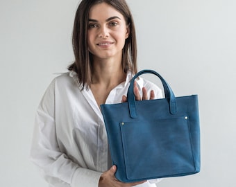 May include: A blue leather tote bag with a front pocket and two top handles. The bag is held by a person wearing a white button-down shirt and tan pants. The bag has a rectangular shape and is a solid blue color.