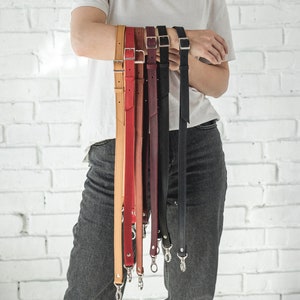 Könnte beinhalten: Eine Auswahl an Leder-Taschenriemen in verschiedenen Farben, darunter Beige, Rot, Burgunder, Schwarz und Braun. Jeder Riemen verfügt über eine silberfarbene Schnalle und Beschläge. Die Riemen werden vor einer weißen Backsteinmauer präsentiert.