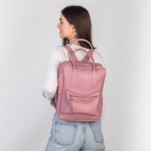 May include: A dusty rose leather backpack with a rectangular shape. The backpack has a front zippered pocket, a top handle, and adjustable shoulder straps. The bag is worn by a person wearing a white long-sleeve shirt and blue jeans.