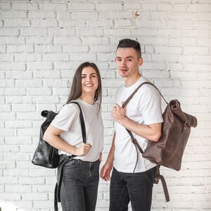 May include: Two people are standing in front of a white brick wall. They are both wearing white t-shirts and carrying brown leather backpacks.