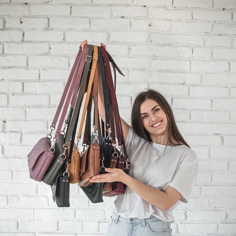 Pode incluir: Uma cole&ccedil;&atilde;o de bolsas crossbody de couro em v&aacute;rias cores, incluindo roxo, cinza, marrom e preto. Cada bolsa tem uma al&ccedil;a longa ajust&aacute;vel e ferragens prateadas. As bolsas s&atilde;o exibidas contra uma parede de tijolos brancos.