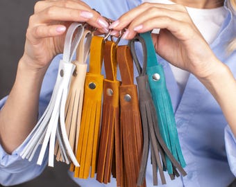 May include: A collection of leather tassel keychains in various colors, including white, beige, yellow, brown, gray, and teal. Each keychain features a metal rivet and a split ring for attaching keys. The tassels are made of fringed leather.