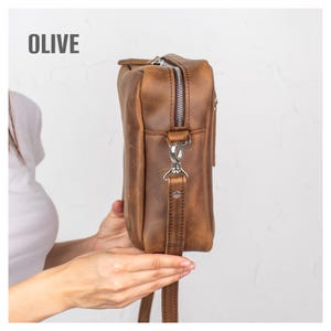 May include: Olive-colored leather crossbody bag with a silver zipper and hardware. The bag has a long, adjustable strap and a rectangular shape. The word "OLIVE" is printed in white at the top. The bag is held by a person.