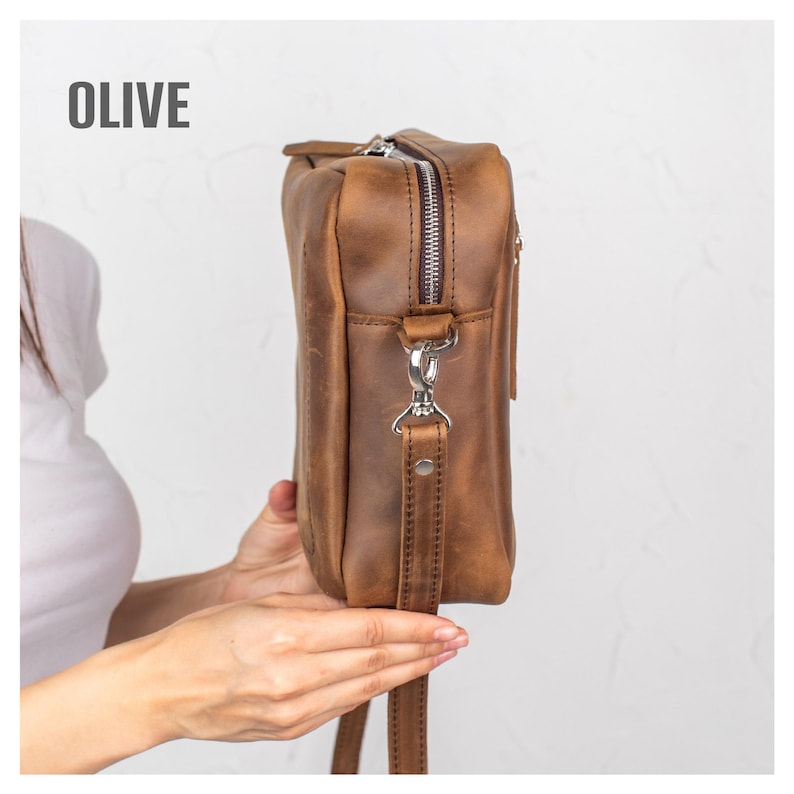May include: Olive-colored leather crossbody bag with a silver zipper and hardware. The bag has a long, adjustable strap and a rectangular shape. The word "OLIVE" is printed in white at the top. The bag is held by a person.
