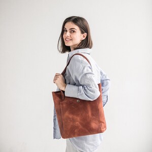 May include: A woman wearing a light blue striped shirt carries a brown leather tote bag with a single snap closure.