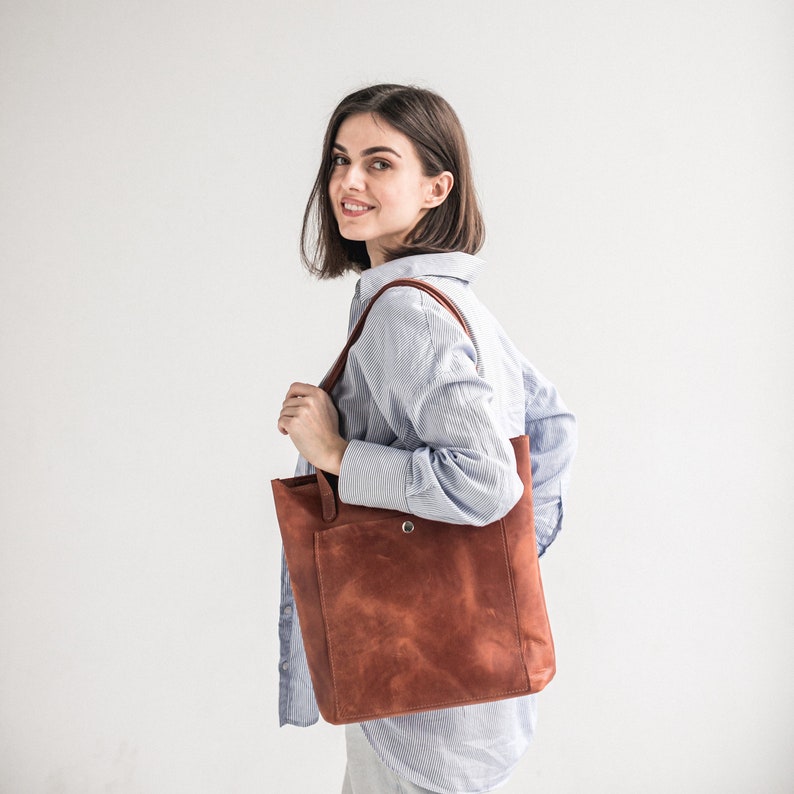 May include: A woman wearing a light blue striped shirt carries a brown leather tote bag with a single snap closure.