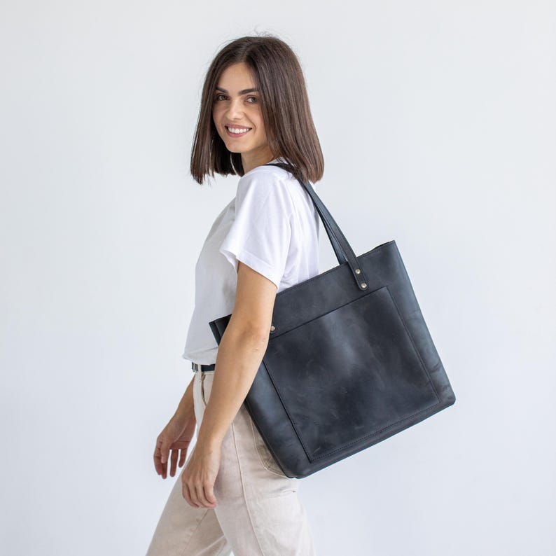 May include: A black leather tote bag with a large front pocket and long shoulder straps. The bag is carried over the shoulder. The bag is a solid black color and appears to be made of high-quality leather.