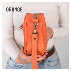 May include: Orange leather crossbody bag with a silver zipper and strap hardware. The bag has a long, detachable strap. The word "ORANGE" is printed in white at the top of the image. The bag is held by a person.