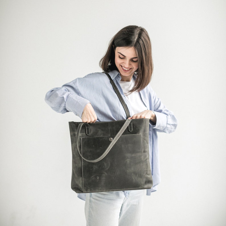 May include: A woman is holding a dark gray leather tote bag with a zippered pocket and a snap closure. The bag has a long strap that can be worn over the shoulder.