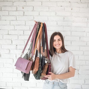 Pu&ograve; includere: Una donna tiene in mano una pila di sette borse a tracolla in pelle di diversi colori. Le borse sono di diverse dimensioni e stili, e sono tutte realizzate in pelle di alta qualit&agrave;.