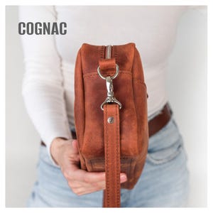 May include: Cognac-colored leather bag with a silver zipper and hardware. The bag has a wrist strap and a rectangular shape. The word "COGNAC" is printed in white at the top of the image.