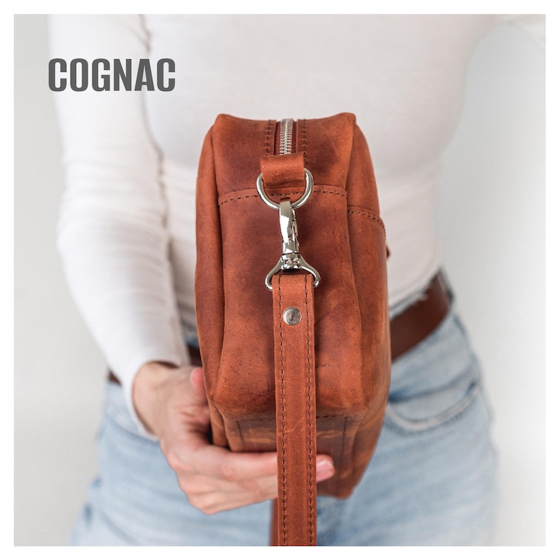 May include: Cognac-colored leather bag with a silver zipper and hardware. The bag has a wrist strap and a rectangular shape. The word "COGNAC" is printed in white at the top of the image.