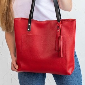 Pode incluir: Uma bolsa tote vermelha brilhante com superfície texturizada, apresentando duas alças pretas presas com rebites metálicos prateados. A bolsa inclui um bolso frontal grande e uma borla decorativa de couro vermelho pendurada numa alça.