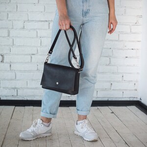 Pu&ograve; includere: Una borsa a tracolla in pelle nera con chiusura a lembo e fibbia argentata. La borsa &egrave; indossata da una persona che indossa jeans blu chiaro e scarpe da ginnastica bianche.