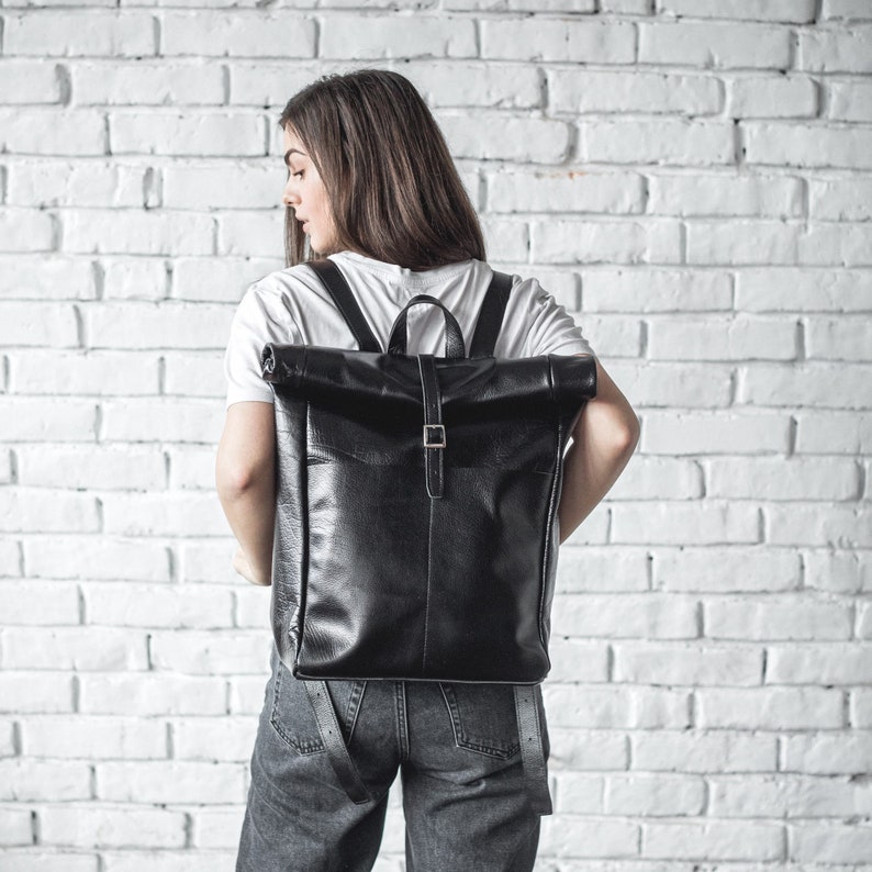 May include: A black leather backpack with a roll-top closure and a buckle strap. The backpack is worn by a person standing in front of a white brick wall.