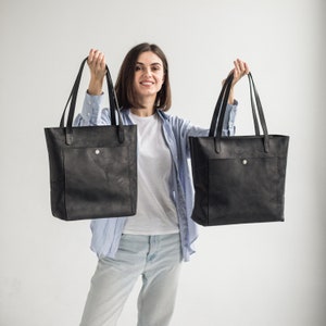 May include: A woman is holding two black leather tote bags with a single pocket on the front of each bag.