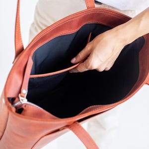 Op de afbeelding: Een roodbruine leren tote bag met een zwart interieur. De tas heeft een ritssluiting en een klein vak aan de buitenkant. De handvatten zijn lang genoeg om over de schouder te dragen. De tas is open en onthult de binnenkant.