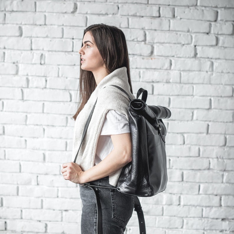 Op de afbeelding: Een zwarte leren roll-top rugzak met een getextureerd oppervlak, een handvat aan de bovenkant en verstelbare schouderbanden. De tas heeft een strak ontwerp met een veilig gespsluitingssysteem voor een functionele opbergoplossing.