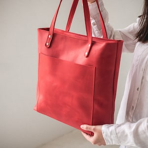 May include: A bright red leather tote bag featuring a large rectangular front pocket, two long shoulder straps attached with silver-toned metal rivets, and a clean, structured design with visible stitching along the edges and pocket seams.