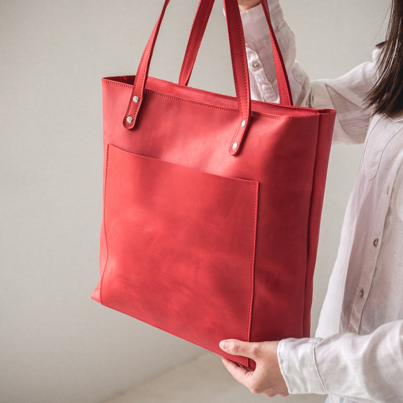 May include: A bright red leather tote bag featuring a large rectangular front pocket, two long shoulder straps attached with silver-toned metal rivets, and a clean, structured design with visible stitching along the edges and pocket seams.