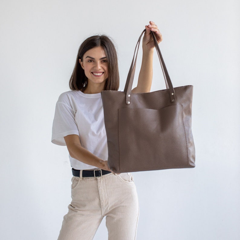 May include: A woman is holding a brown leather tote bag with a front pocket. The bag has a simple design and is made of a soft, textured leather.