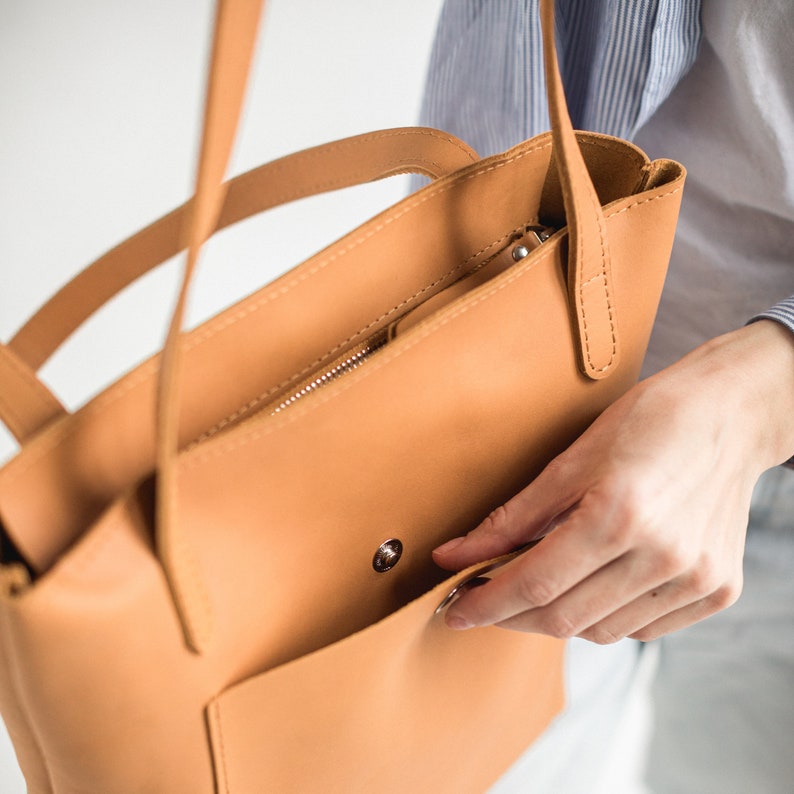 May include: A tan leather tote bag with a zippered pocket and a front pocket. The bag has a top handle and a shoulder strap.