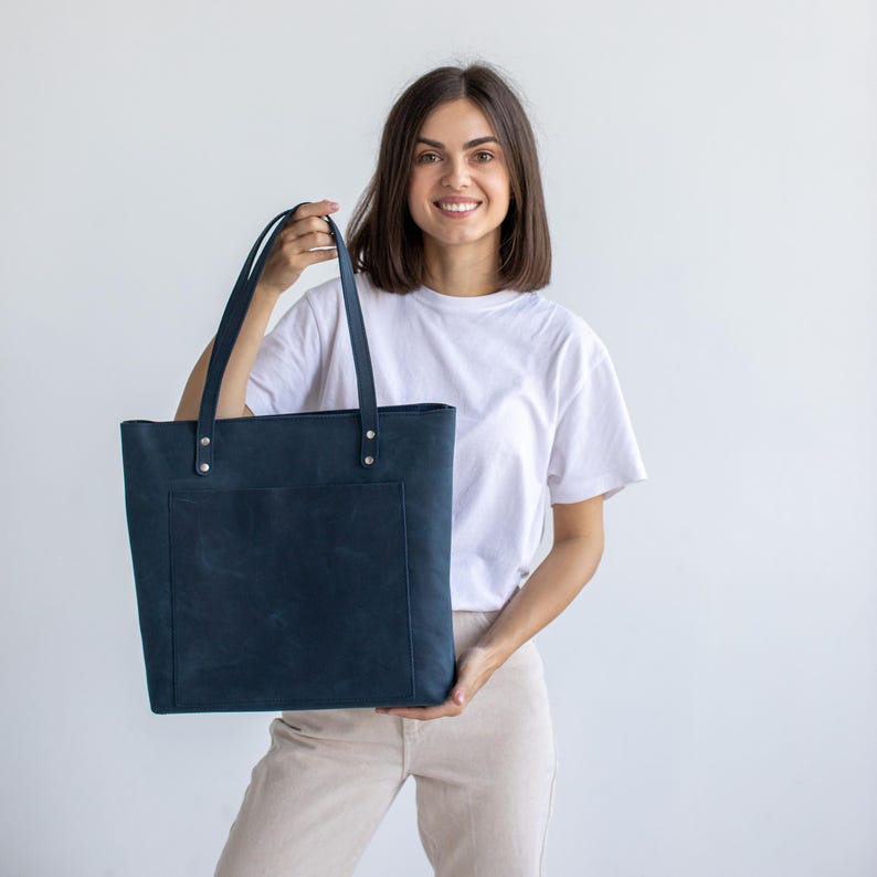 May include: A dark navy blue leather tote bag with a large rectangular front pocket, two long shoulder straps, and silver-toned metal rivets securing the straps to the top edge of the bag, featuring a smooth matte finish and clean lines.