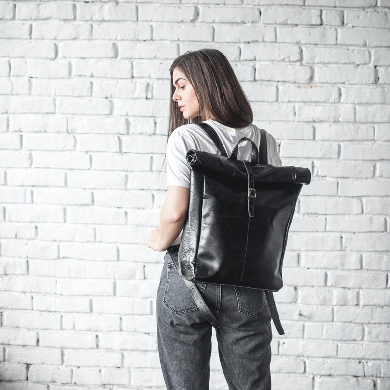 Op de afbeelding: Een zwarte leren rugzak met rolsluiting, voorzien van een verticale riem met metalen gesp, een handvat aan de bovenzijde en verstelbare schouderbanden. De tas heeft een gladde, halfglanzende afwerking en een rechthoekige vorm met een omgevouwen bovenkant.