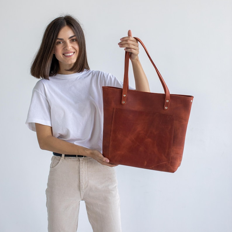 A brown leather tote bag with a front pocket and two handles. The bag is held up by a person. The bag has a rectangular shape and is made of a textured leather material. The bag is a reddish-brown color.