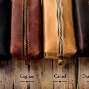 May include: Four leather pencil cases in black, cognac, camel, and dark brown. Each case has a brass zipper and a leather pull tab. The names of the colors are written below each case on a wooden surface.