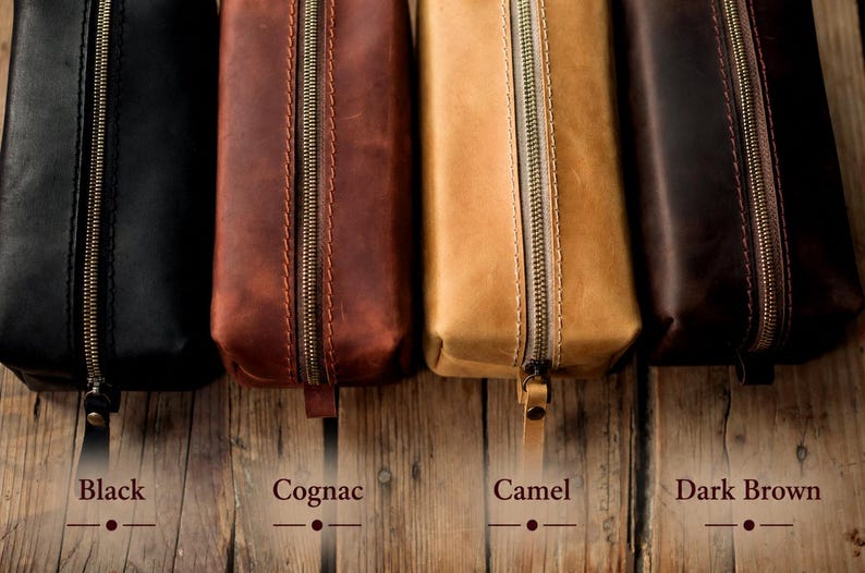 May include: Four leather pencil cases in black, cognac, camel, and dark brown. Each case has a brass zipper and a leather pull tab. The names of the colors are written below each case on a wooden surface.