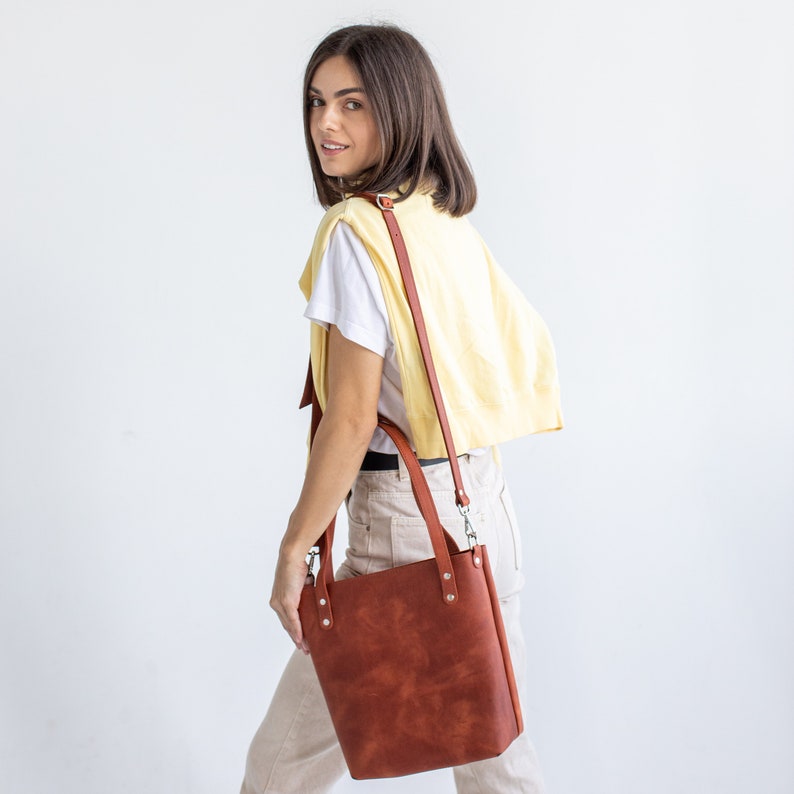 May include: A structured, rectangular leather tote bag in a rich, reddish-brown hue. The bag features two sturdy leather handles and a long, adjustable shoulder strap attached with silver-toned metal hardware and rivets on the front panel.