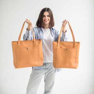 May include: A woman is holding two large, tan leather tote bags with a front pocket. The bags have a simple design and are made of a high-quality material.