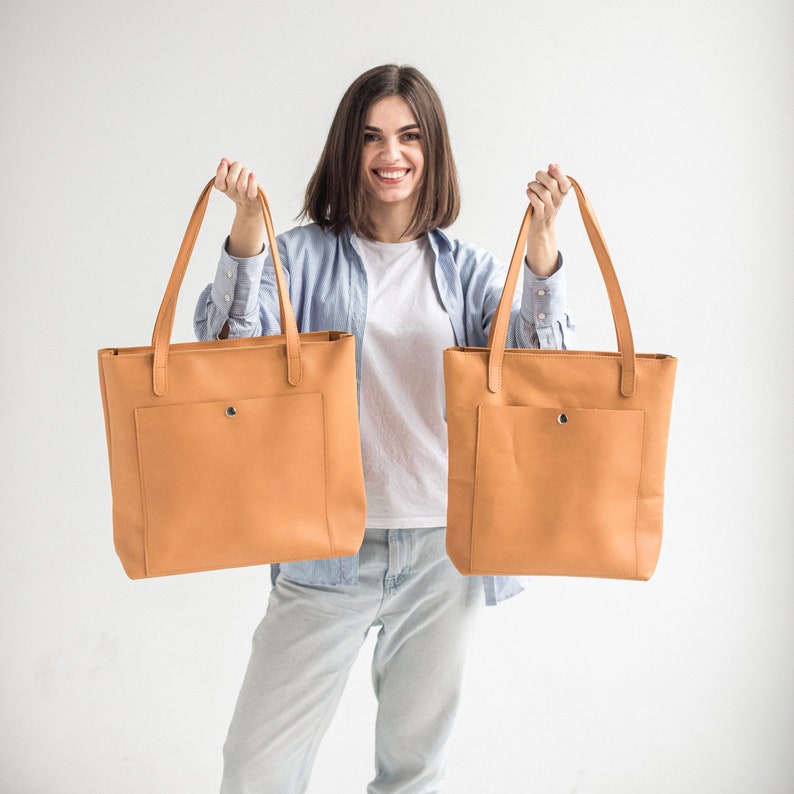 May include: A woman is holding two large, tan leather tote bags with a front pocket. The bags have a simple design and are made of a high-quality material.