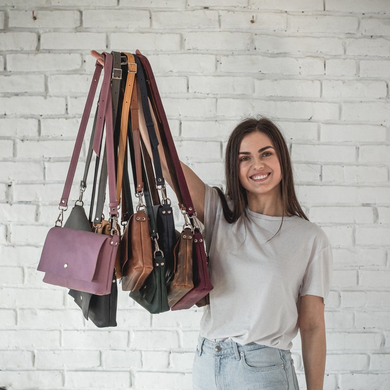 Pode incluir: Uma cole&ccedil;&atilde;o de bolsas crossbody de couro em v&aacute;rias cores, incluindo roxo, preto, marrom e azul marinho. Cada bolsa apresenta uma al&ccedil;a longa e ajust&aacute;vel e um fecho de aba. As bolsas s&atilde;o exibidas contra uma parede de tijolos brancos.