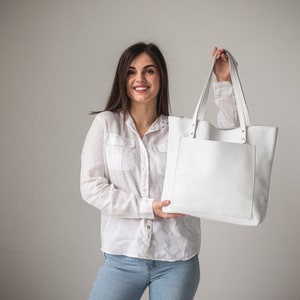 May include: A woman is holding a white leather tote bag with a front pocket. The bag has a simple design and is made of high-quality leather.