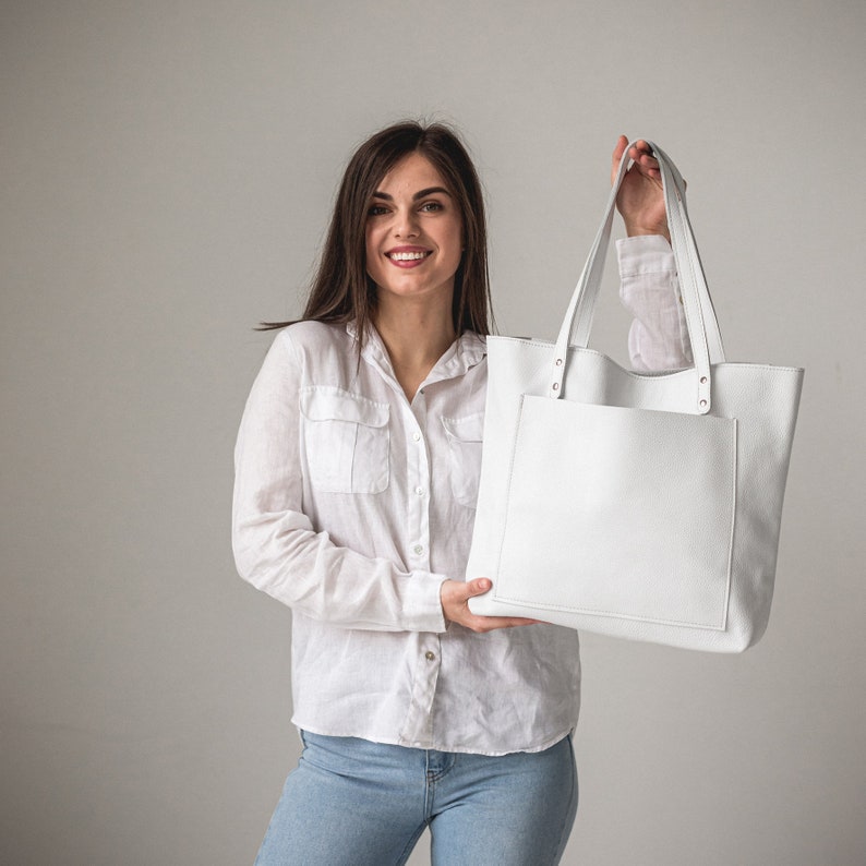 May include: A woman is holding a white leather tote bag with a front pocket. The bag has a simple design and is made of high-quality leather.