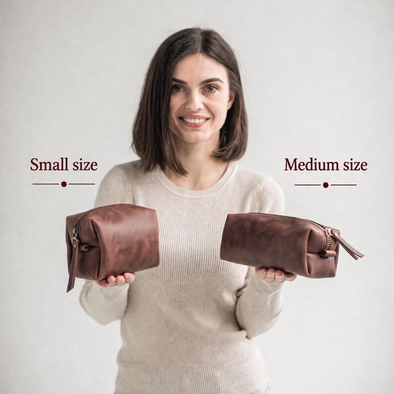 May include: Two brown leather toiletry bags, a small and a medium size, are displayed. The small bag is held in the left hand, and the medium bag is held in the right hand. Both bags have a zipper closure and a leather pull.
