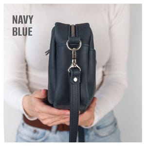Pode incluir: Bolsa transversal de couro azul marinho com z&iacute;per e ferragens prateadas. A bolsa tem uma al&ccedil;a longa e ajust&aacute;vel e um pequeno bolso lateral. O texto "NAVY BLUE" est&aacute; impresso no canto superior esquerdo.