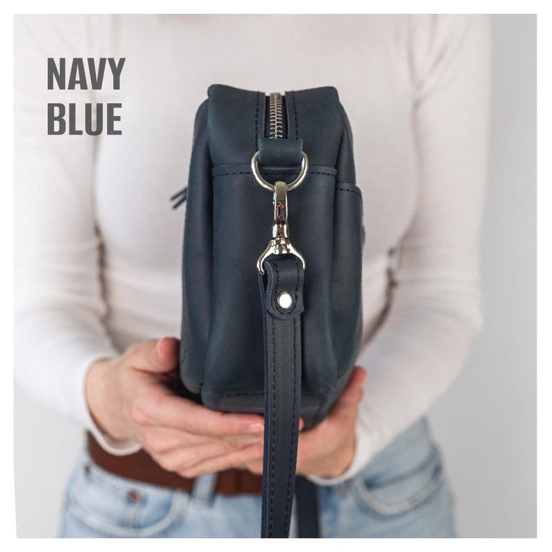 Pode incluir: Bolsa transversal de couro azul marinho com z&iacute;per e ferragens prateadas. A bolsa tem uma al&ccedil;a longa e ajust&aacute;vel e um pequeno bolso lateral. O texto "NAVY BLUE" est&aacute; impresso no canto superior esquerdo.