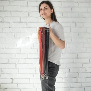 Pode incluir: Uma cole&ccedil;&atilde;o de al&ccedil;as de couro para bolsas em v&aacute;rias cores, incluindo bege, vermelho, bord&ocirc; e preto. Cada al&ccedil;a tem uma fivela ou fechos de metal para fixa&ccedil;&atilde;o. As al&ccedil;as s&atilde;o mantidas verticalmente, mostrando seus diferentes tons e texturas.