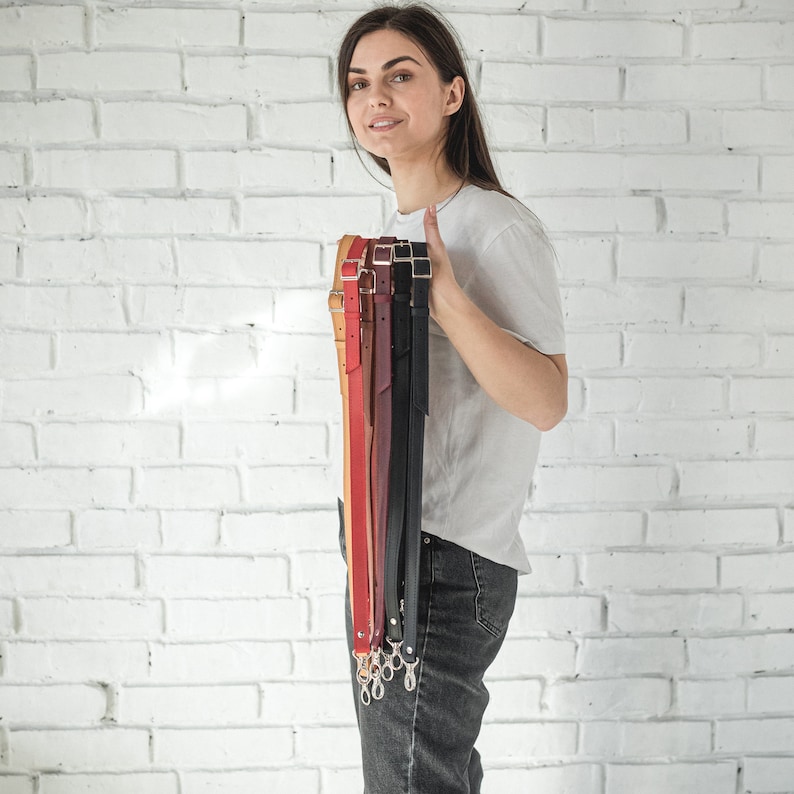 Pode incluir: Uma cole&ccedil;&atilde;o de al&ccedil;as de couro para bolsas em v&aacute;rias cores, incluindo bege, vermelho, bord&ocirc; e preto. Cada al&ccedil;a tem uma fivela ou fechos de metal para fixa&ccedil;&atilde;o. As al&ccedil;as s&atilde;o mantidas verticalmente, mostrando seus diferentes tons e texturas.