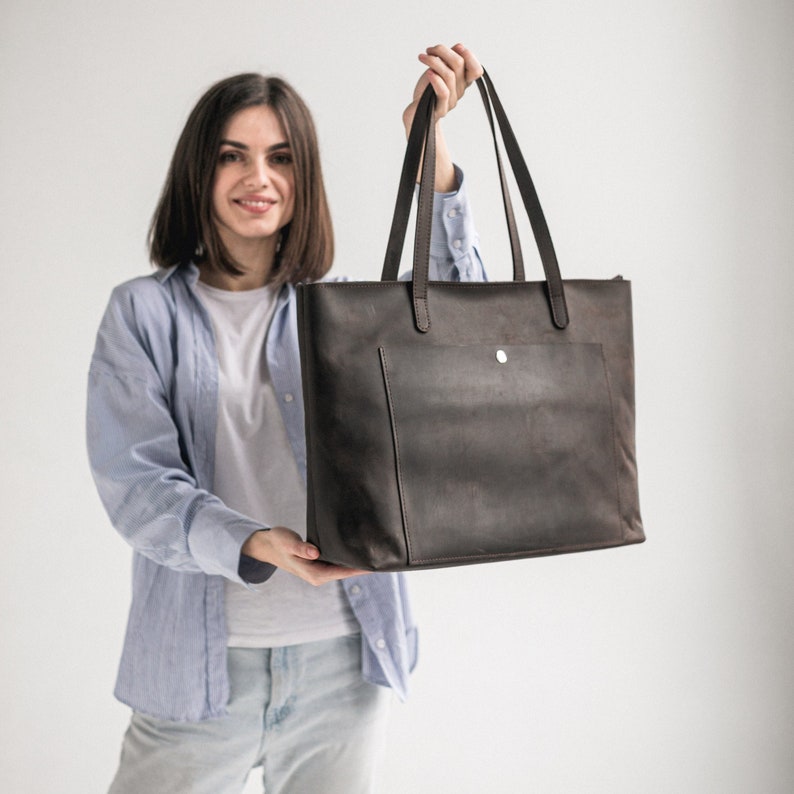 May include: A brown leather tote bag with a zipper closure and a front pocket. The bag has a single strap and is held by a person.