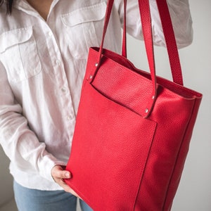 May include: A bright red leather tote bag with a front pocket. The bag has a top handle and two shoulder straps.