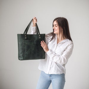 May include: A dark forest green leather tote bag with two shoulder straps, a large rectangular front pocket, and metallic rivets securing the handles to the main body of the bag. The bag has a smooth, matte finish with visible natural texture.