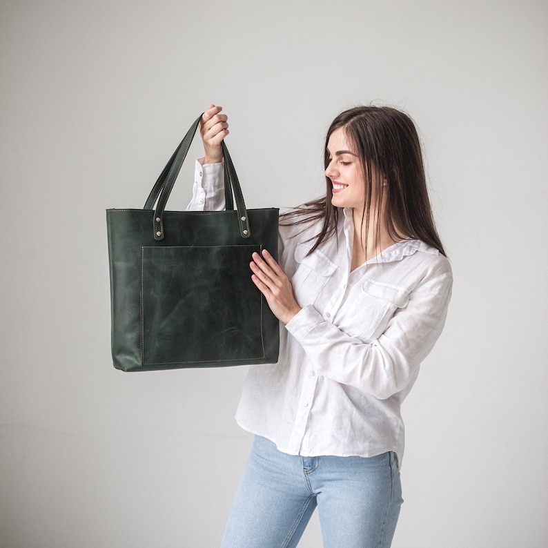 May include: A dark forest green leather tote bag with two shoulder straps, a large rectangular front pocket, and metallic rivets securing the handles to the main body of the bag. The bag has a smooth, matte finish with visible natural texture.