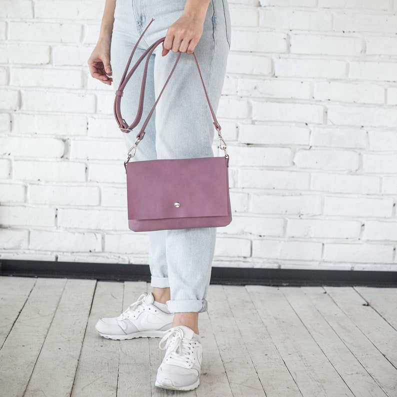 Pu&ograve; includere: Una borsa a tracolla in pelle color malva con una fibbia argentata e una lunga tracolla regolabile. La borsa &egrave; indossata da una persona che indossa jeans blu chiaro e scarpe da ginnastica bianche.