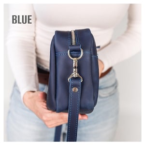 May include: A blue leather crossbody bag with a silver zipper and strap hardware. The bag has a long, adjustable strap and the word "BLUE" is printed in the upper left corner. The bag is held by a person.